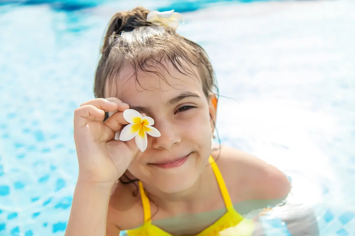 Flicka i gul baddräkt i en pool, ler och håller en blomma framför ena ögat.